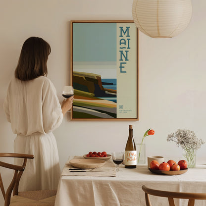 A woman in a beige dress holds a glass of wine while admiring a colorful Maine poster on the wall. A dining table with fruits and wine is in the.
