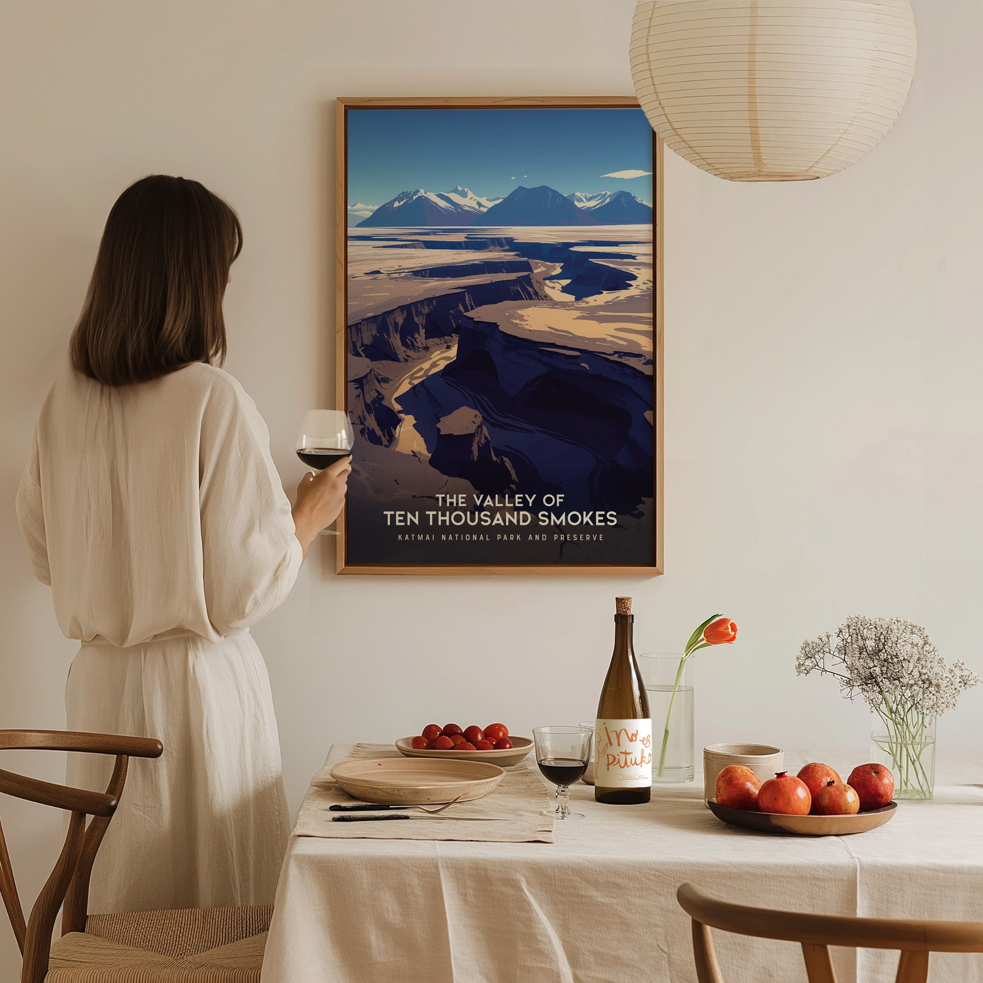 A woman in a white dress holds a glass of wine, admiring a Valley of Ten Thousand Smokes poster on the wall above a dining table with fruit and flowers.