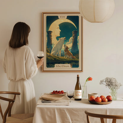 A woman in a white dress holds a wine glass, admiring a framed The Moon tarot poster. The table in front is set with fruit and a wine bottle.