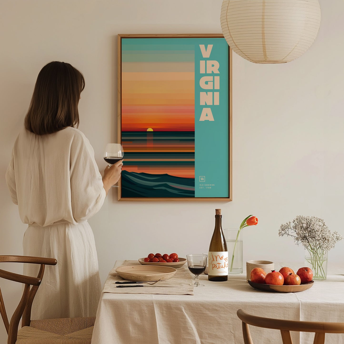 A person in a white dress holds a glass of wine, admiring a colorful Virginia poster on the wall. A table with fruit, wine, and flowers is in the foreground.