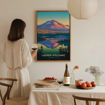 A woman in a white dress holds a glass of wine, admiring a Lassen Volcanic National Park poster. The table features a wine bottle, apples, and a vase with flowers.