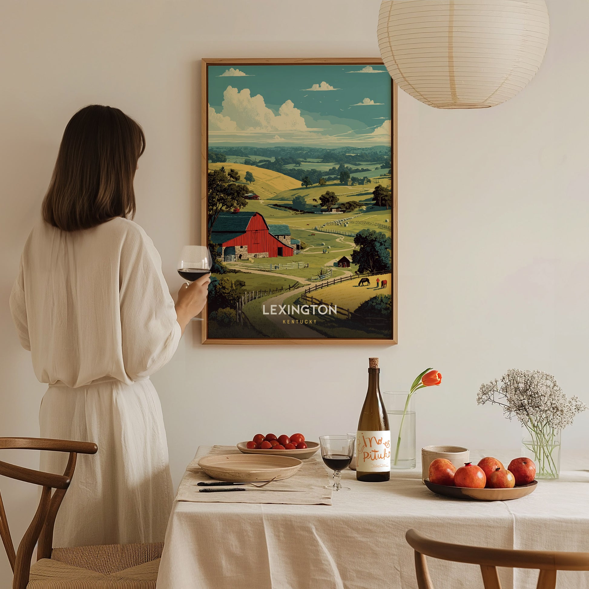 A woman in a white dress stands holding a glass of wine, gazing at a framed poster depicting a serene landscape with rolling hills, a red barn, and grazing animals.