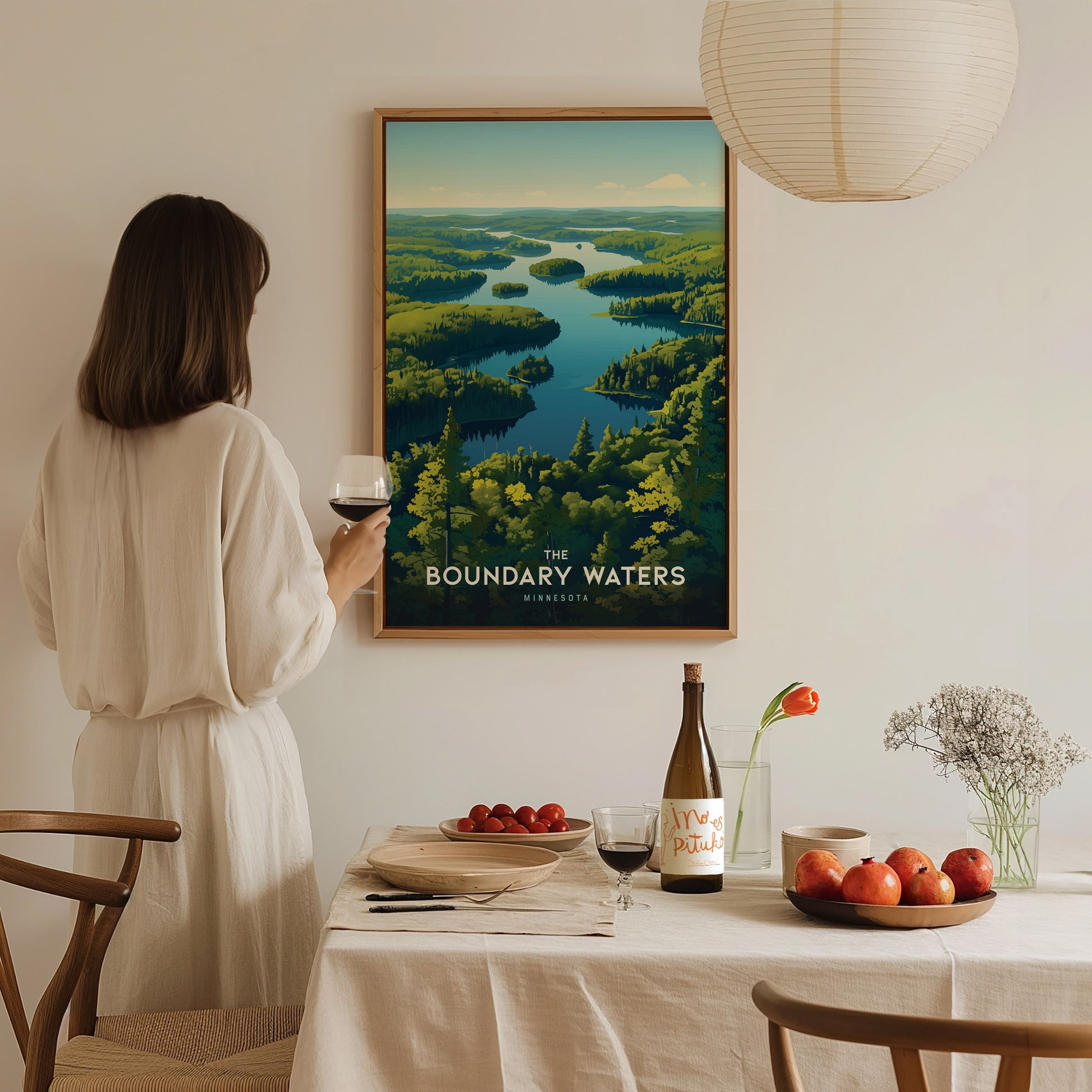 A woman in a white dress holds a glass of wine while admiring a framed poster of Boundary Waters. The table is set with fruit, flowers, and a wine bottle.