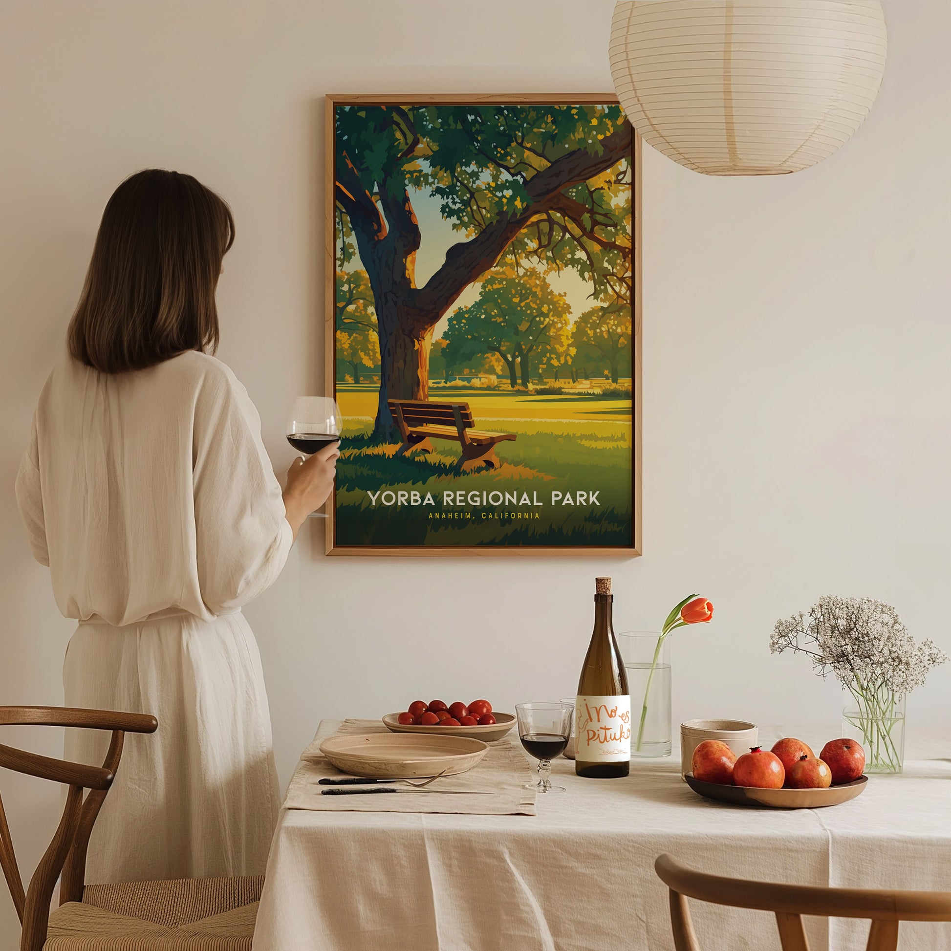 A woman in a white dress holds a glass of wine, admiring a Yorba Regional Park poster. A dining table with fruit and wine is elegantly set beneath.