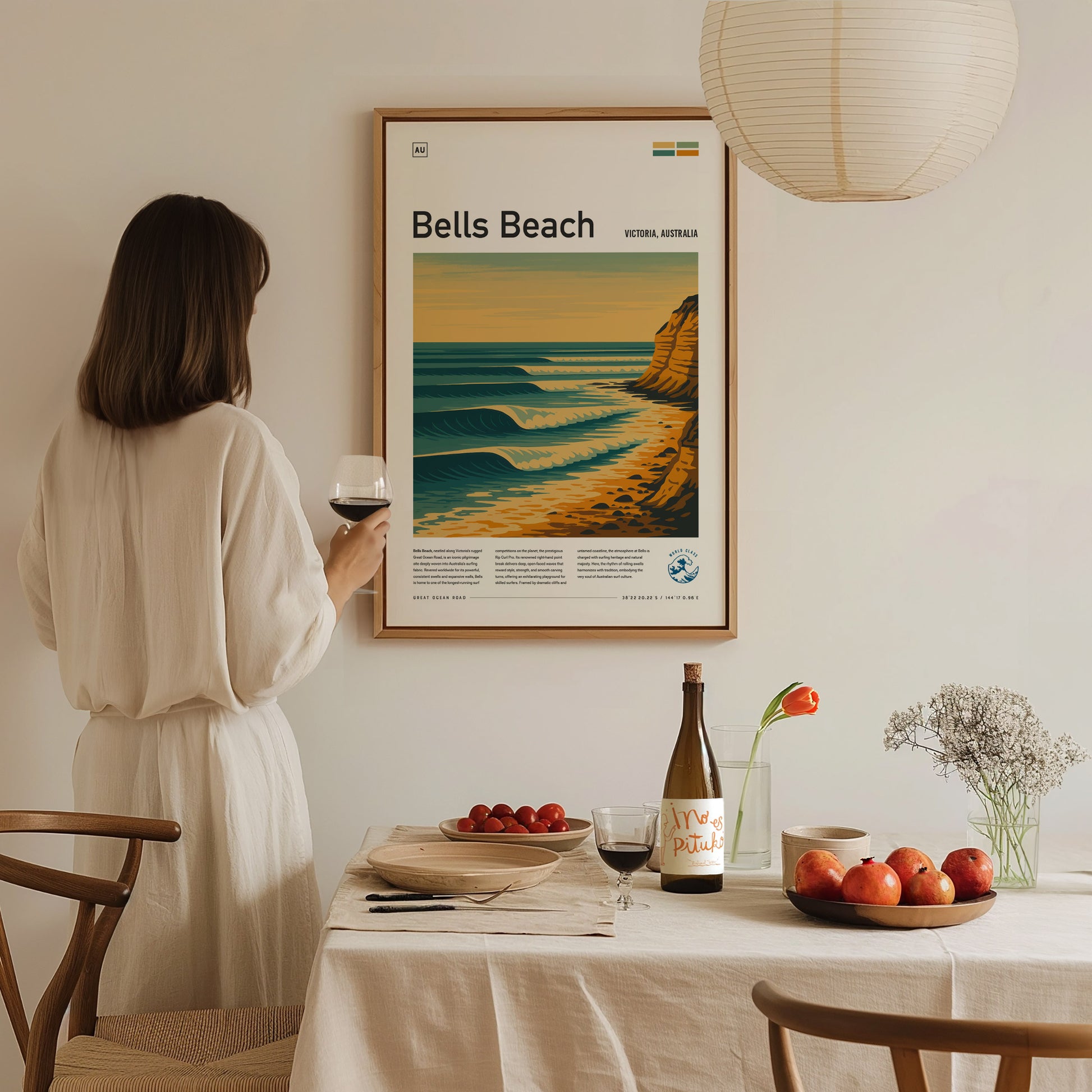 A person in a white dress admires a framed Bells Beach poster on the wall. A table with wine, fruit, and flowers is set in the foreground.