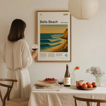 A person in a white dress admires a framed Bells Beach poster on the wall. A table with wine, fruit, and flowers is set in the foreground.