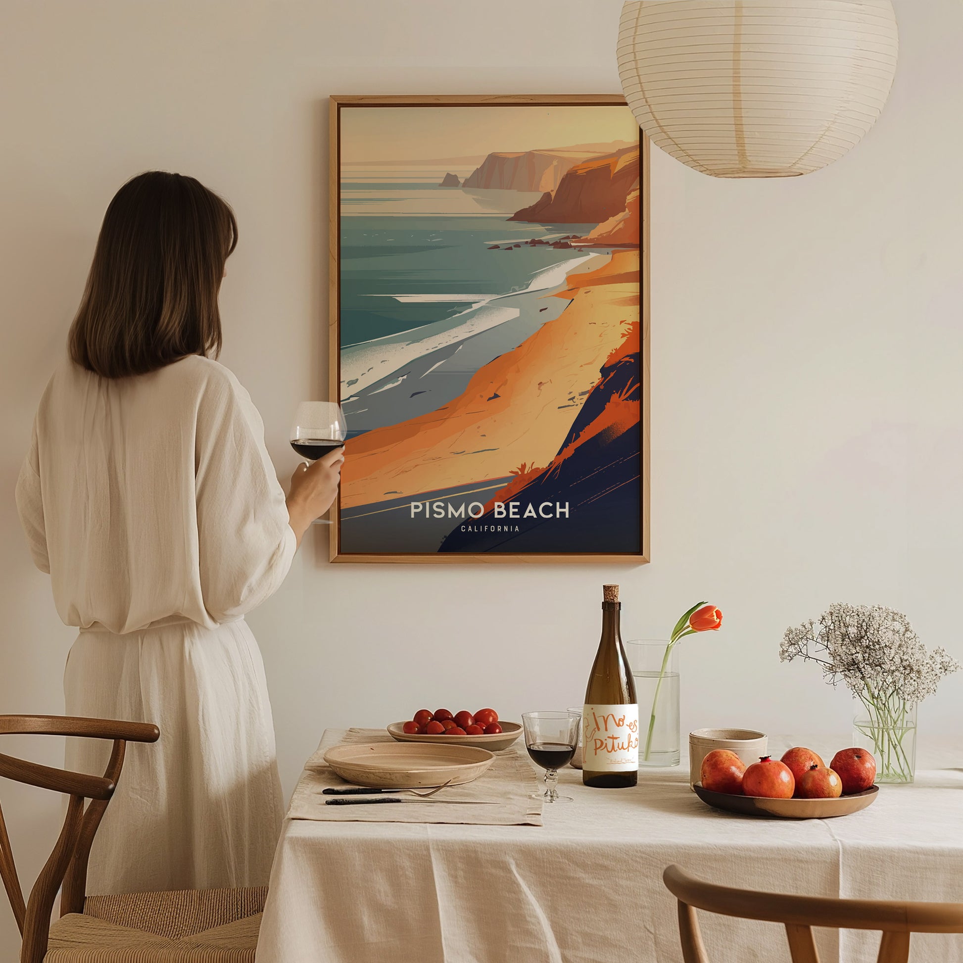 A woman in a white dress admires a framed Pismo Beach poster on the wall, with a table set with wine, fruit, and flowers in the foreground.