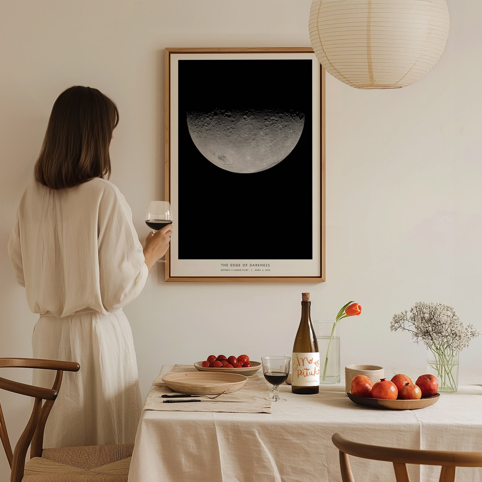 A woman in a light-colored outfit stands holding a glass of wine, gazing at a framed poster of the moon hanging on a wall.