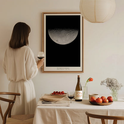 A woman in a light-colored outfit stands holding a glass of wine, gazing at a framed poster of the moon hanging on a wall.