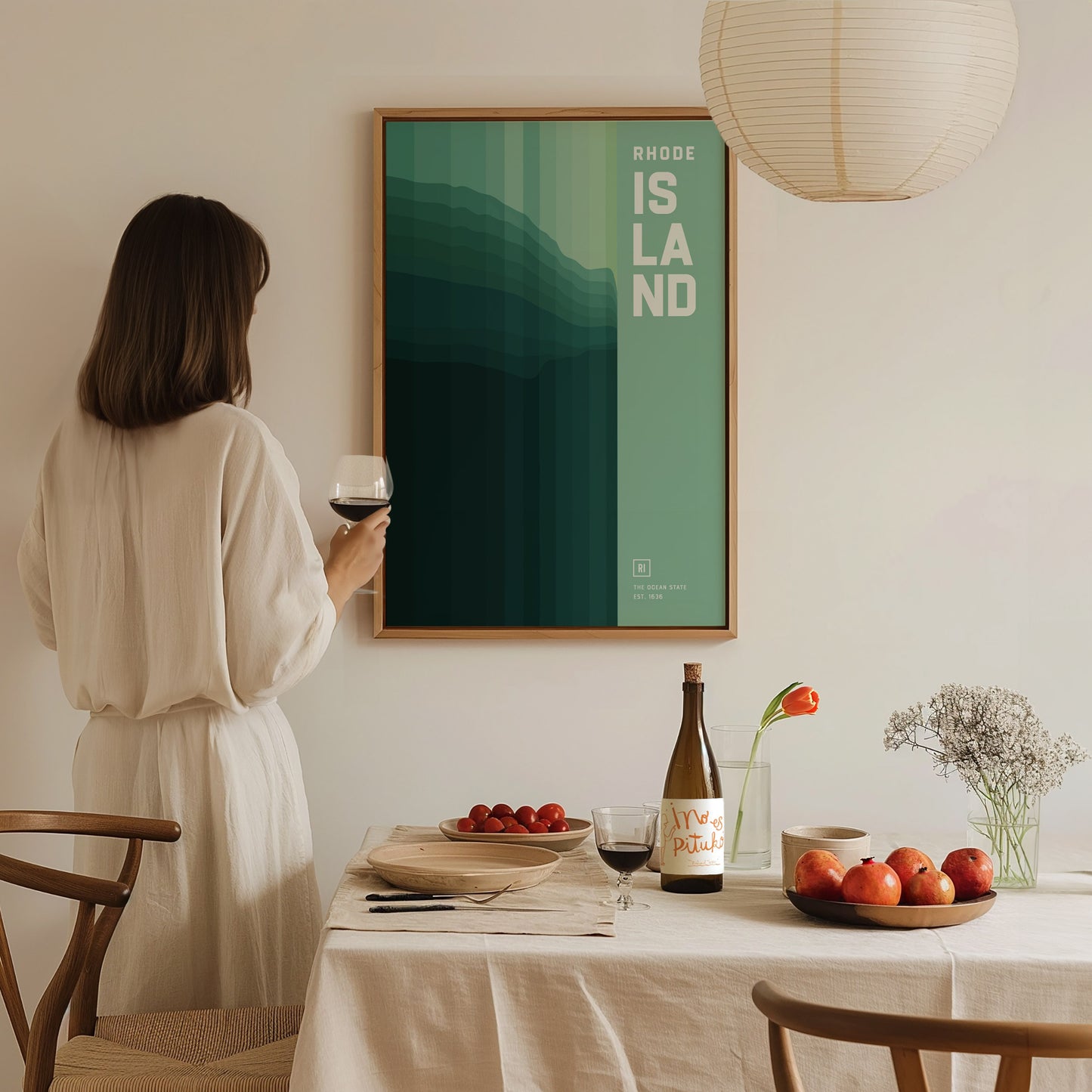 A woman in a white dress holds a wine glass, admiring a framed abstract Rhode Island poster on the wall. A table with fruit and wine completes the cozy setting.