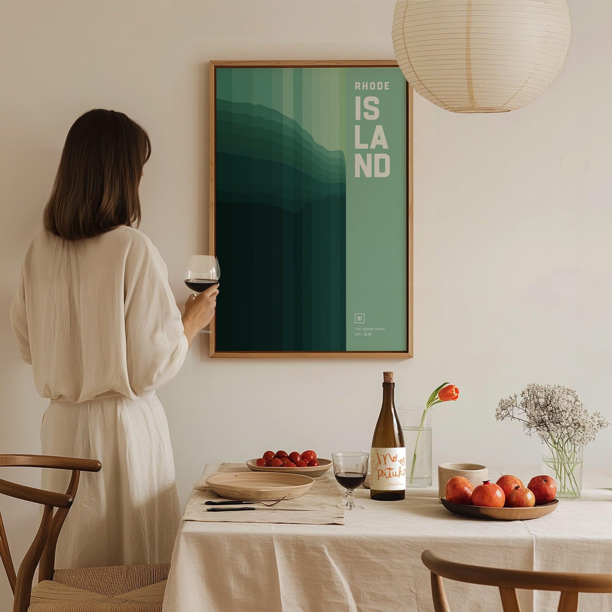 A woman in a white dress holds a wine glass, admiring a framed abstract Rhode Island poster on the wall. A table with fruit and wine completes the cozy setting.