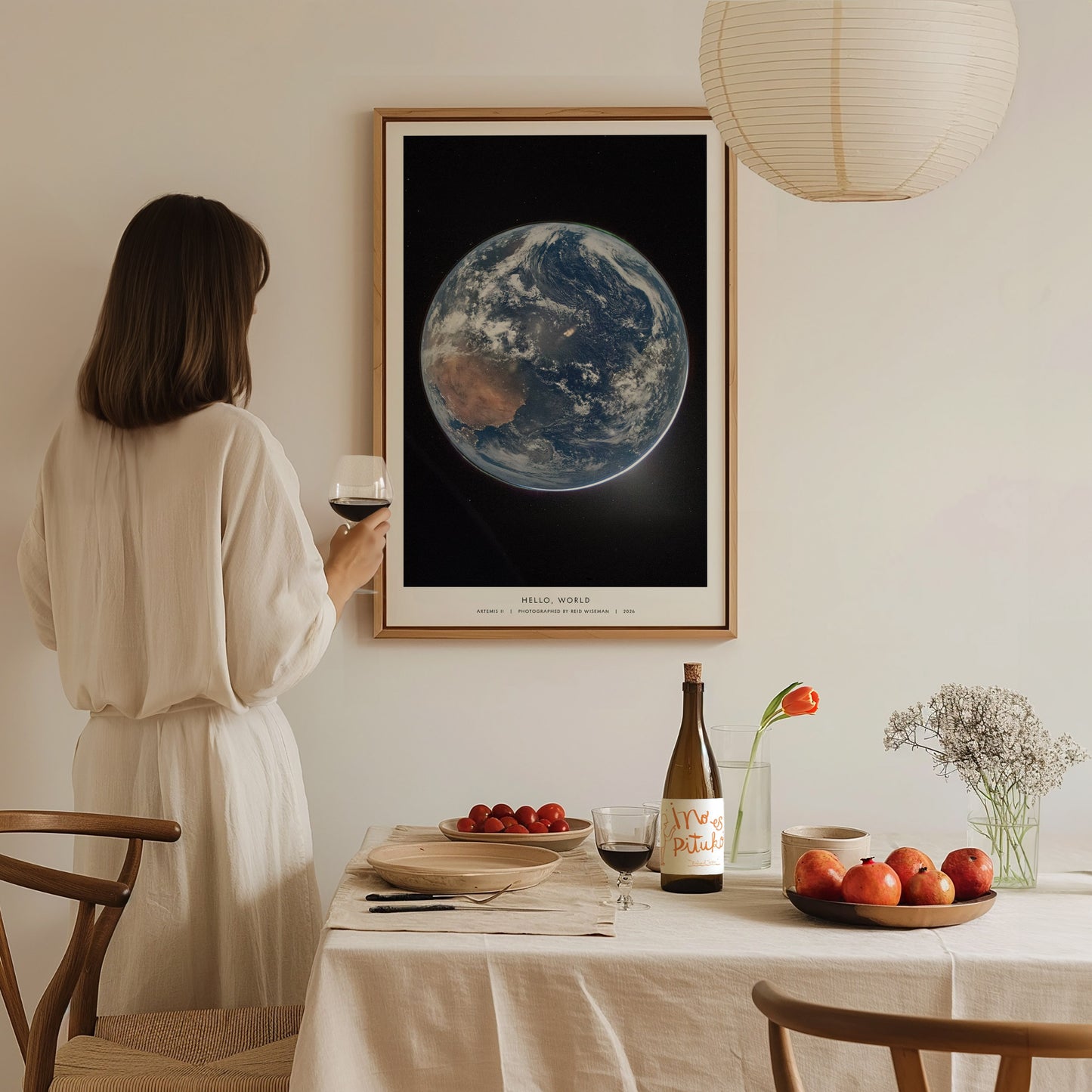 A woman in a white dress holds a glass of wine while admiring a framed poster of Earth from space, featuring vibrant colors and detailed imagery.