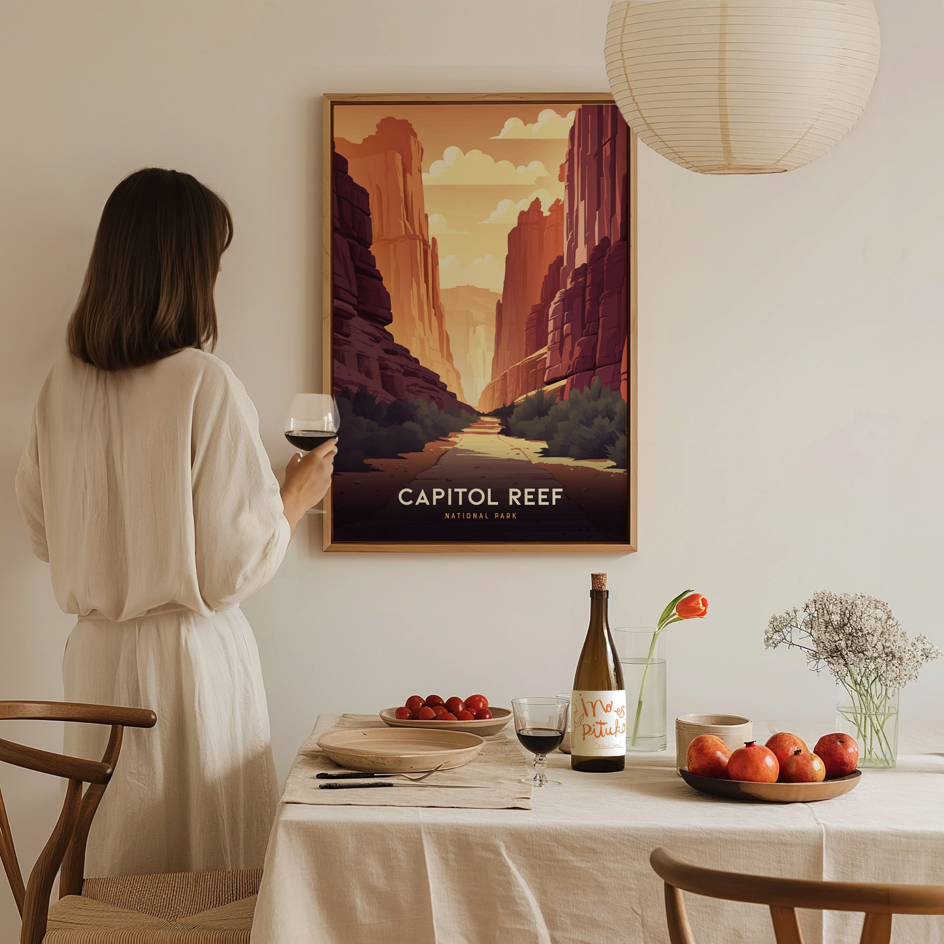 A woman in a white dress holds a glass of wine, admiring a Capitol Reef National Park poster on the wall. The table is set with fruit, wine, and flowers.