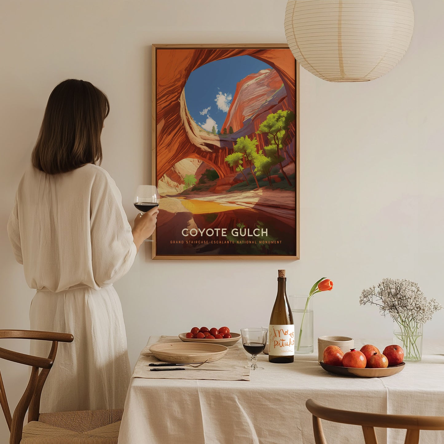 A woman in a white dress holds a glass of wine, admiring a framed Coyote Gulch poster on the wall. A dining table with fruits and wine is in the.