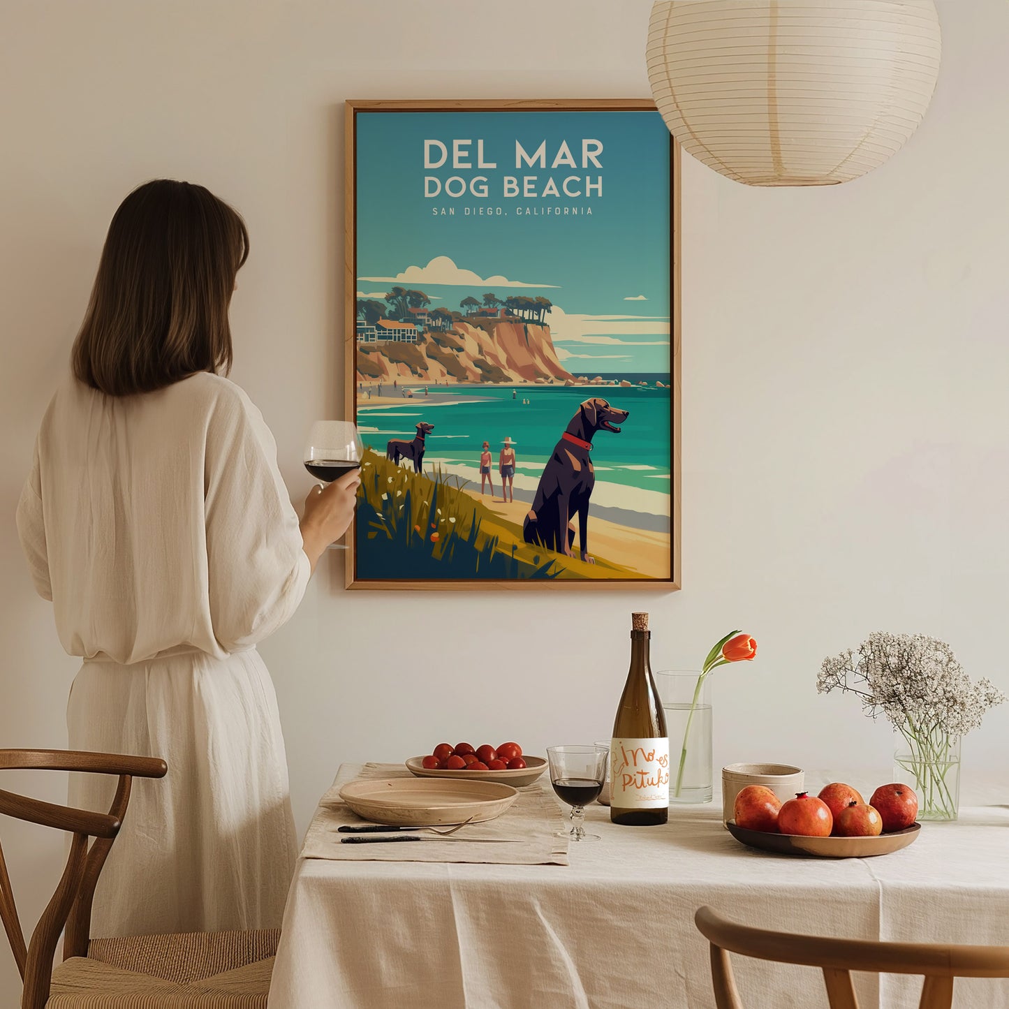 A woman in a white dress admires a Del Mar Dog Beach poster in a cozy dining room, featuring a coastal landscape with dogs and cliffs.