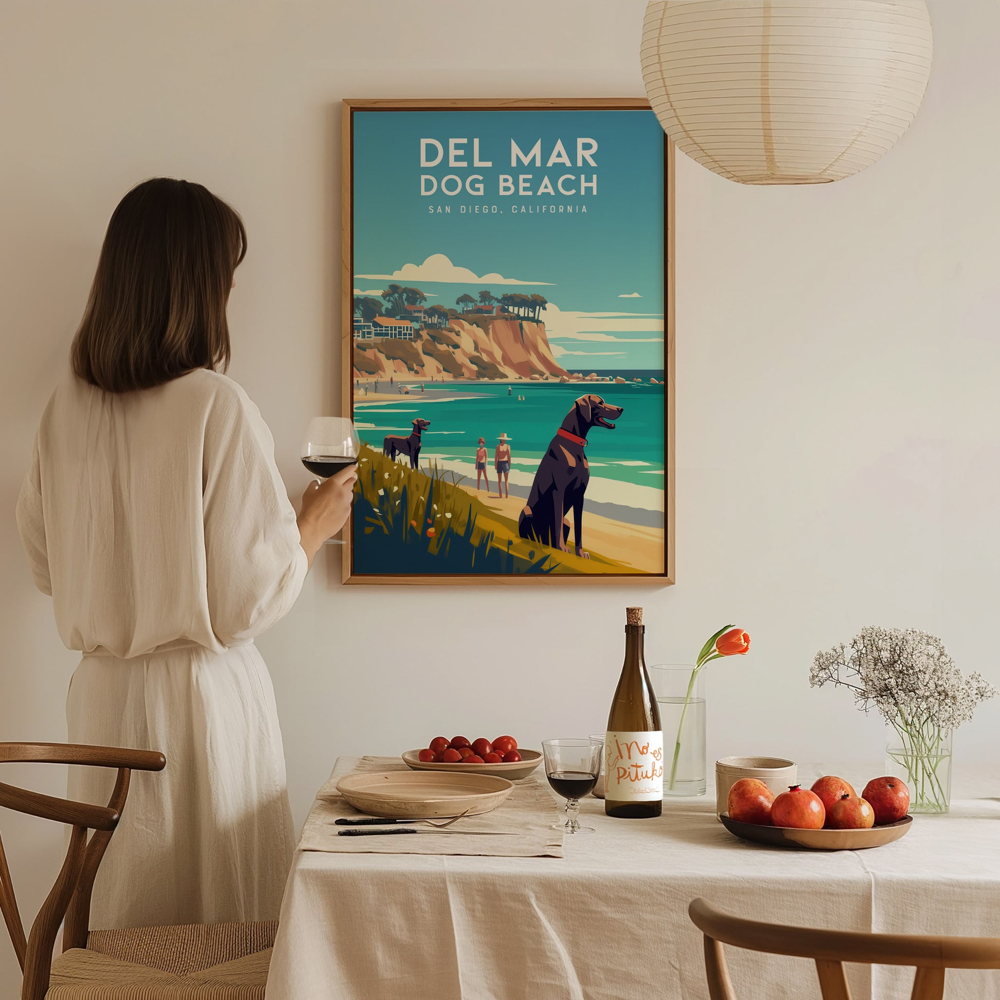 A woman in a white dress admires a Del Mar Dog Beach poster in a cozy dining room, featuring a coastal landscape with dogs and cliffs.