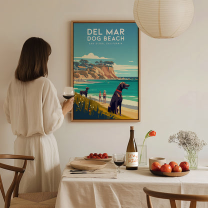 A woman in a white dress admires a Del Mar Dog Beach poster in a cozy dining room, featuring a coastal landscape with dogs and cliffs.