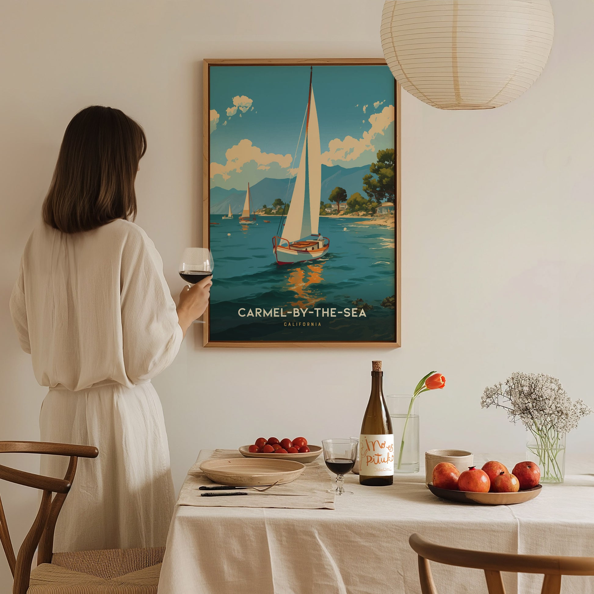 A woman in a white dress admires a Carmel-by-the-Sea sailboat poster on the wall. A table in the foreground is set with wine, fruit, and flowers.