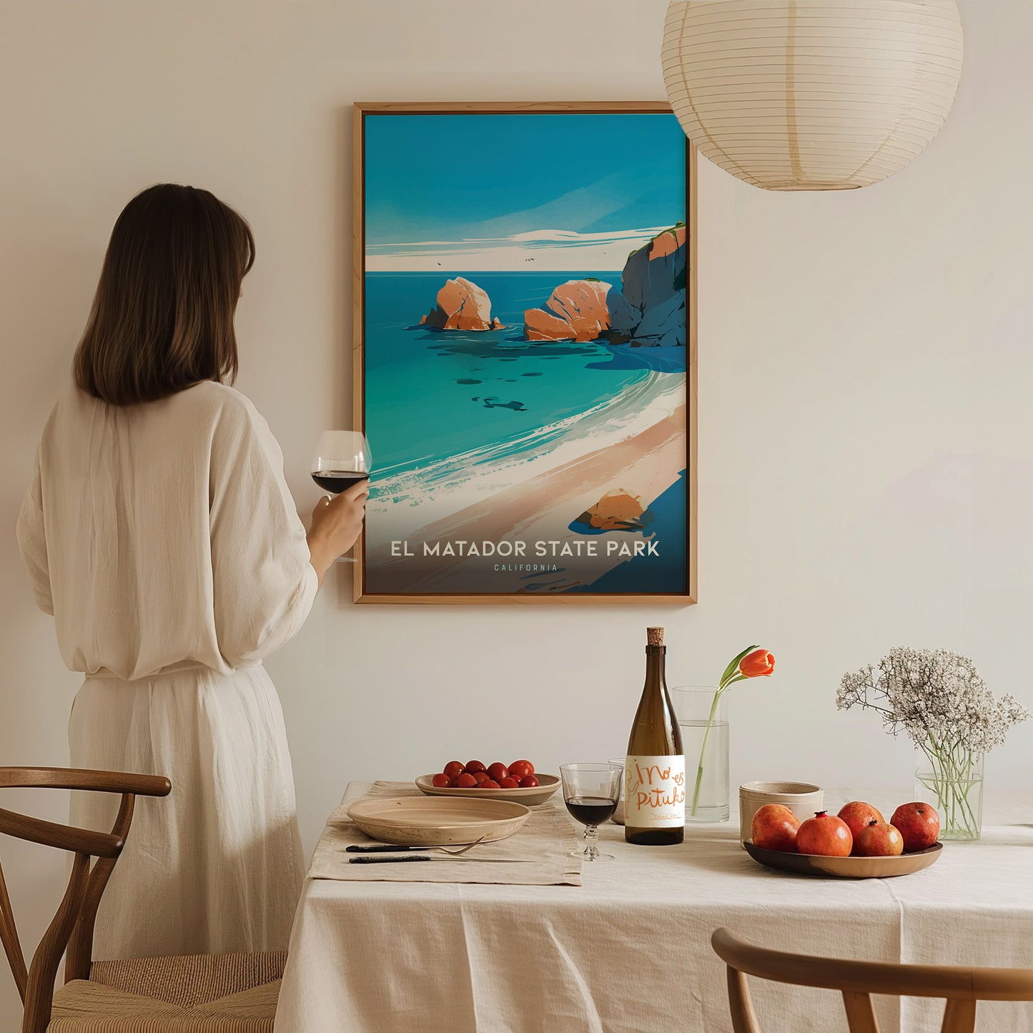 A woman in a white dress holds a glass of wine, admiring a colorful El Matador State Park poster on the wall.