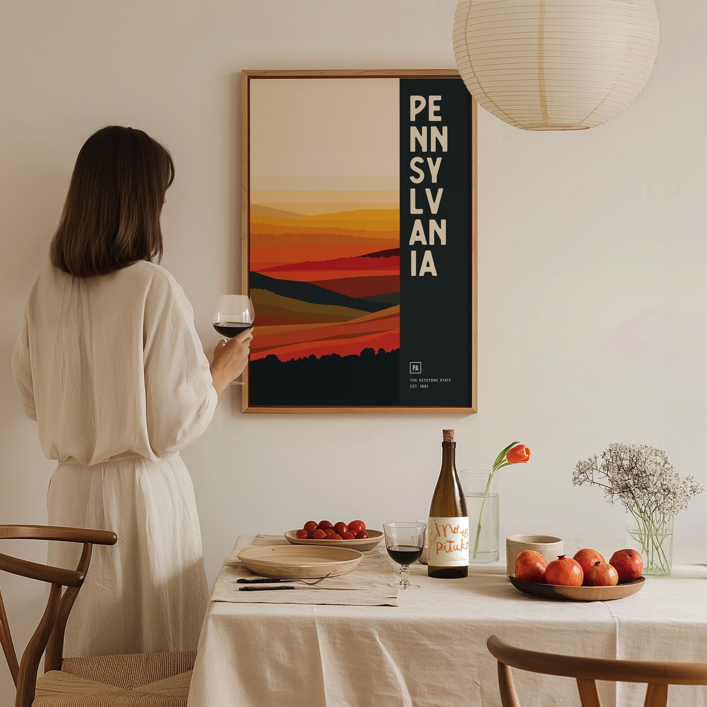 A woman in a white dress holding a glass of wine stands in front of a Pennsylvania poster with abstract landscape art.