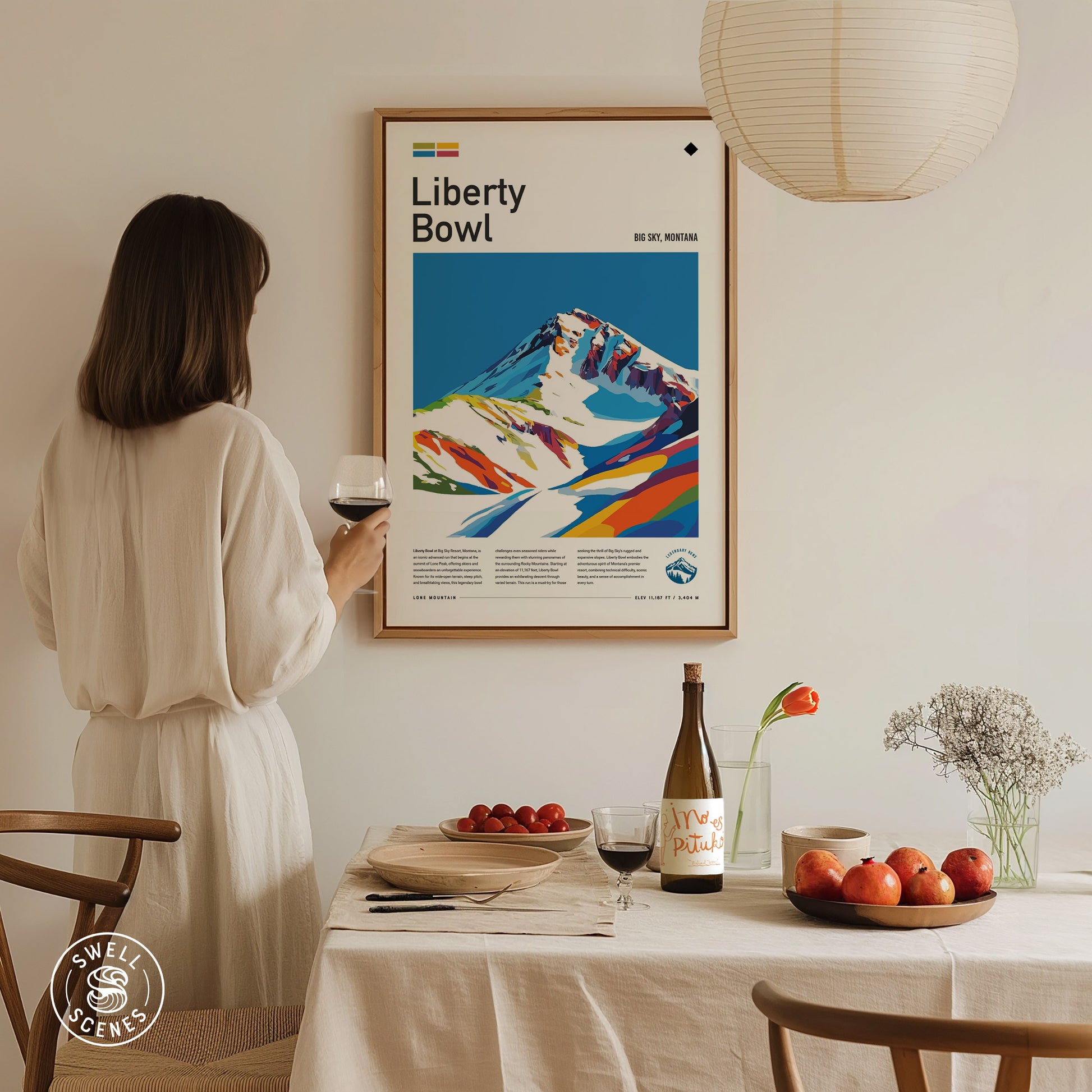 A woman in a white dress holds a glass of wine, admiring a colorful Liberty Bowl Big Sky poster on the wall.