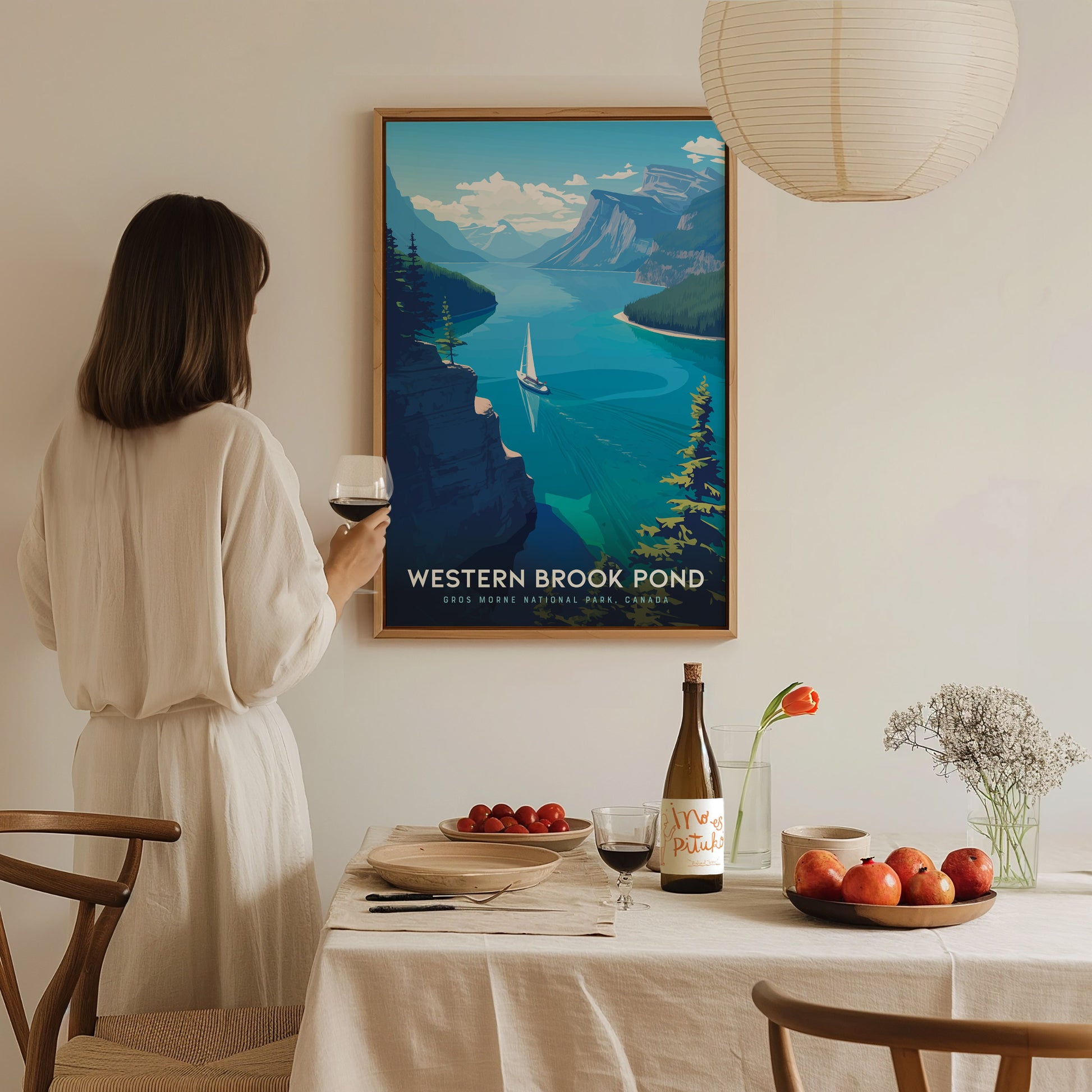 A woman in a white dress holds a glass of wine while looking at a framed Western Brook Pond poster. The dining table is set with fruit, wine, and flowers.