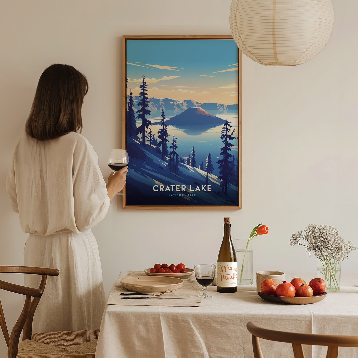 A woman in a white dress admires a framed Crater Lake poster on a wall. A dining table is set with a wine bottle, glass, and a plate of apples.