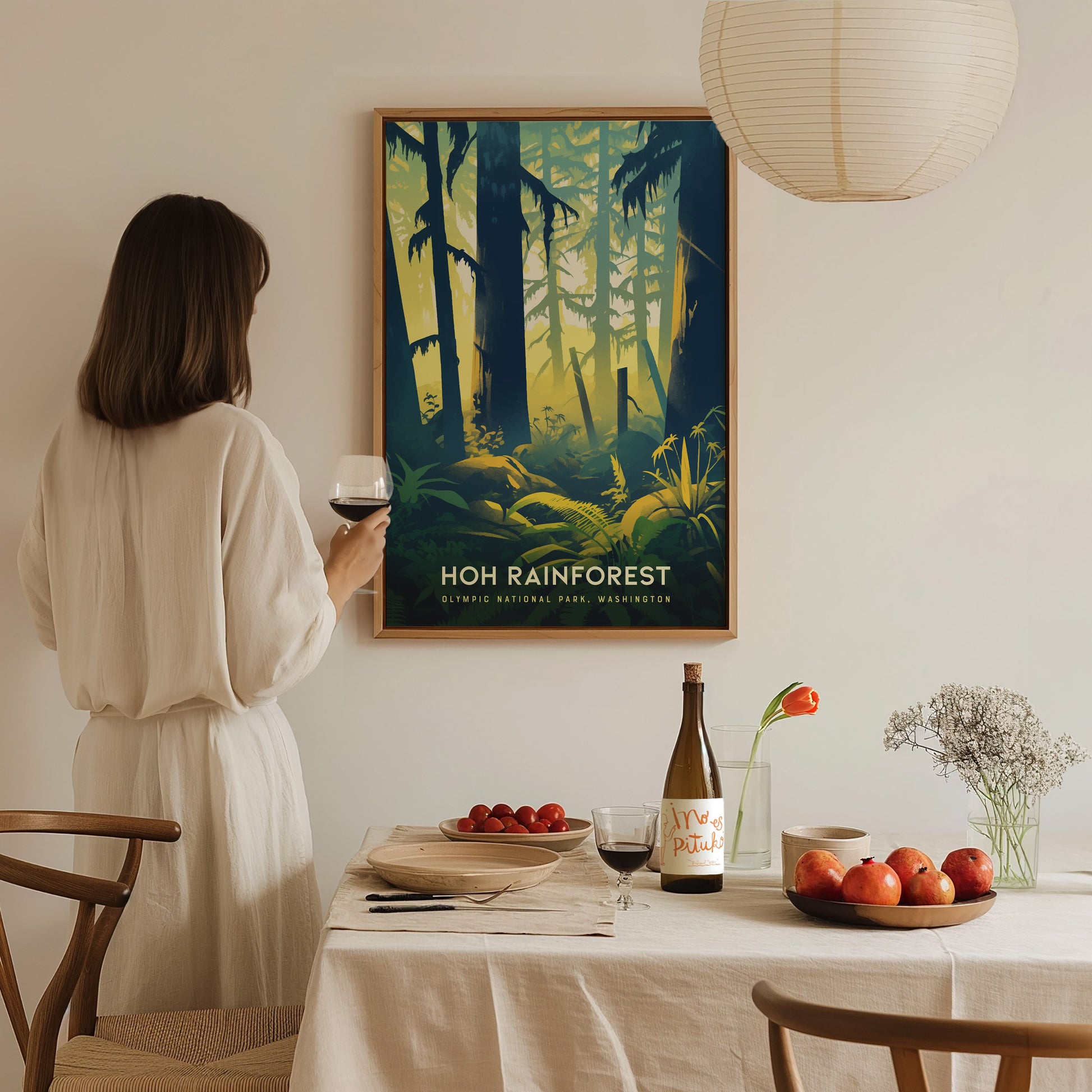 A woman in a white dress holds a wine glass, looking at a Hoh Rainforest poster. A table with fruit, wine, and flowers is in the foreground.