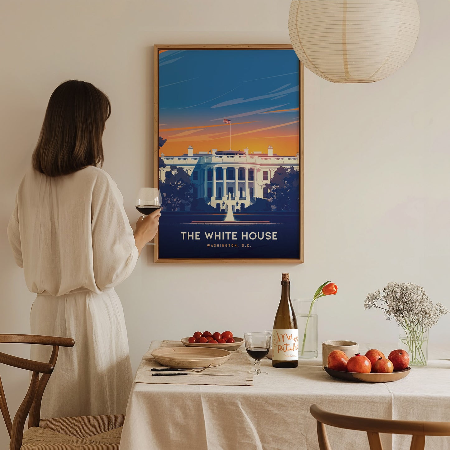 A woman in a white dress holds a glass of wine, gazing at a vibrant White House poster on the wall. A table is set with fruit, wine, and flowers.