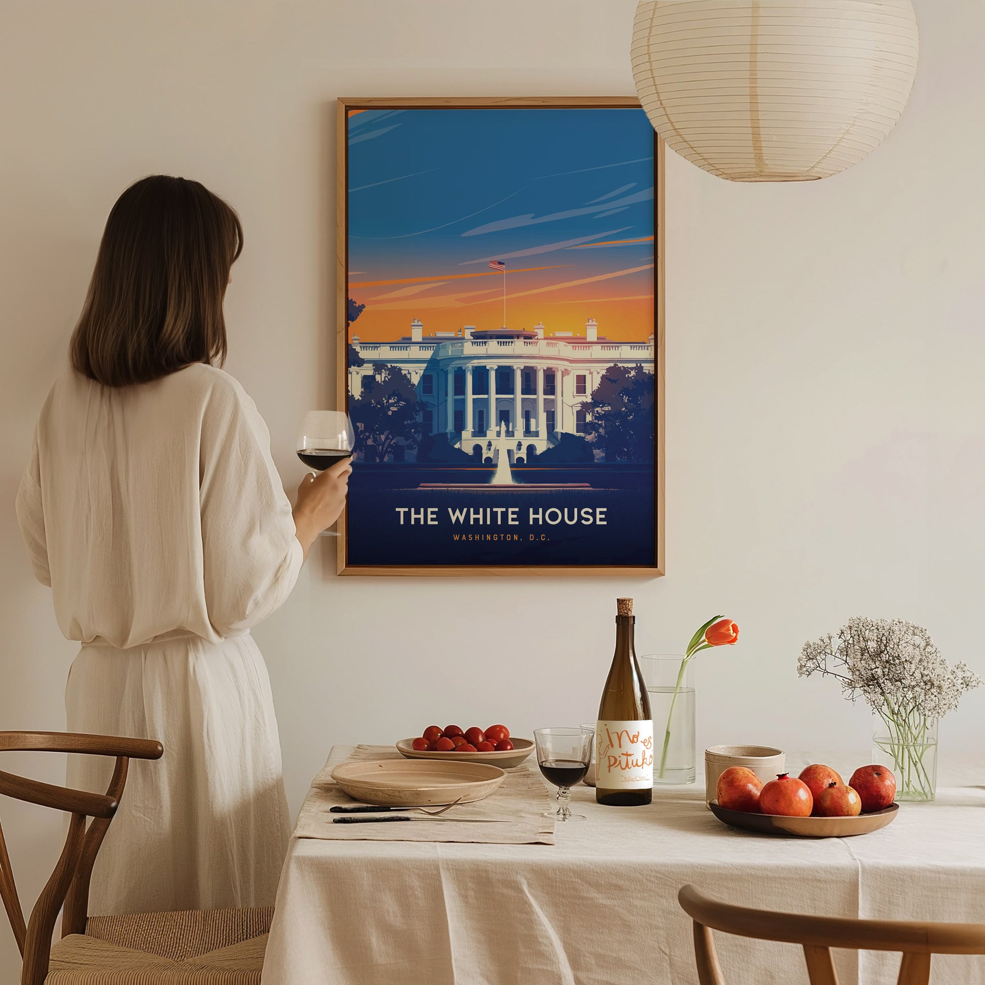 A woman in a white dress holds a glass of wine, gazing at a vibrant White House poster on the wall. A table is set with fruit, wine, and flowers.