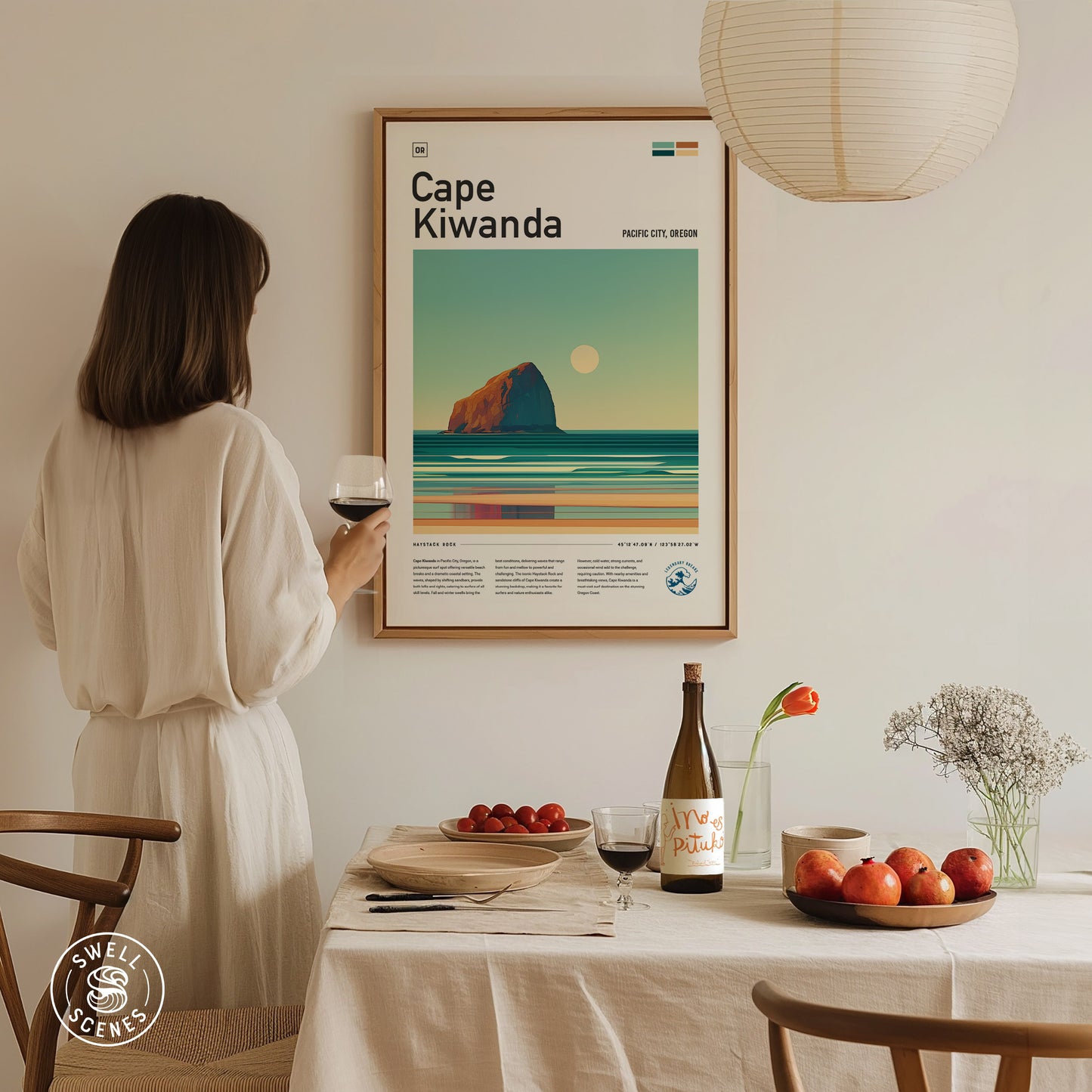 A woman in a white dress holds a glass of wine while admiring a Cape Kiwanda poster on a wall. A table with fruit, wine, and flowers is in the.
