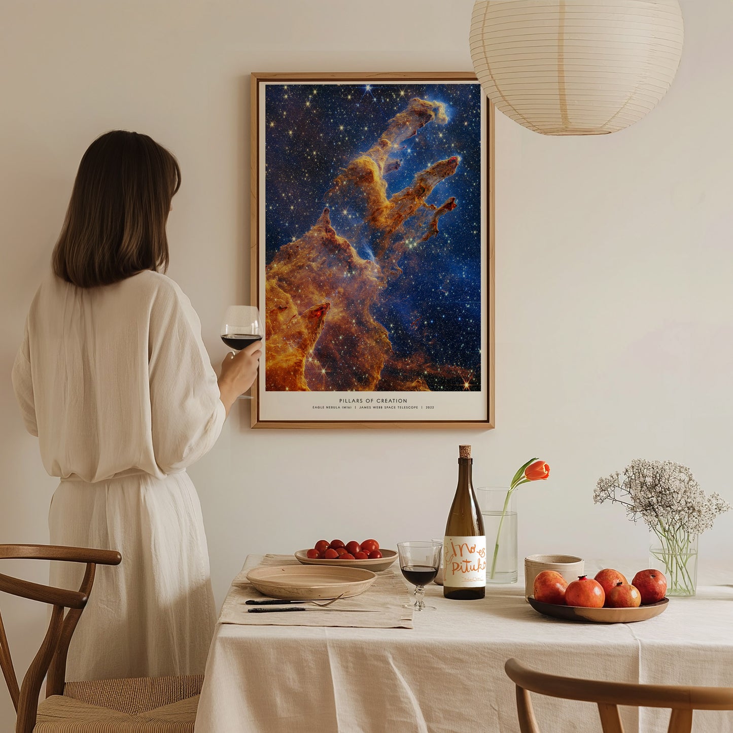 Interior dining scene with a woman standing near a wall-mounted Pillars of Creation JWST poster, framed in natural wood with warm sunlight in the room.