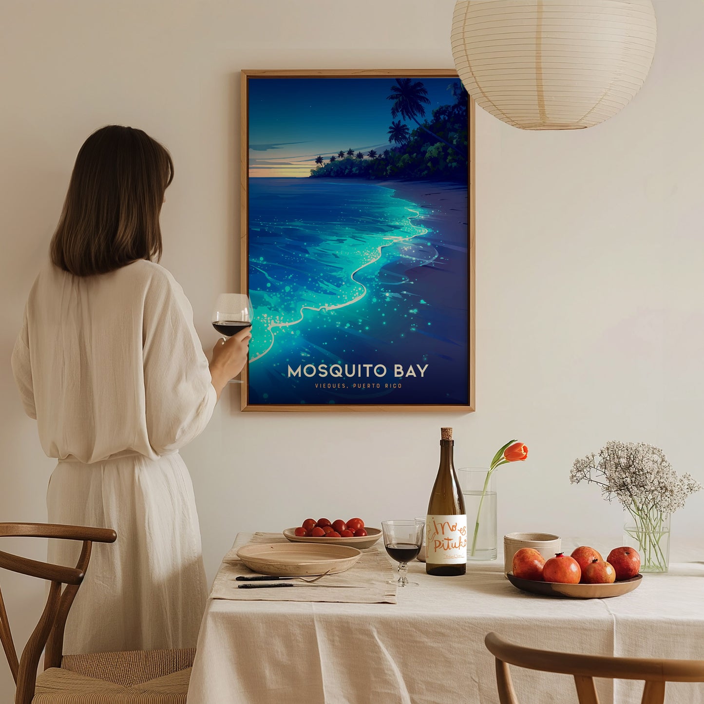 A woman in a light-colored dress stands in a cozy dining area, holding a glass of wine while admiring a framed poster of Mosquito Bay.