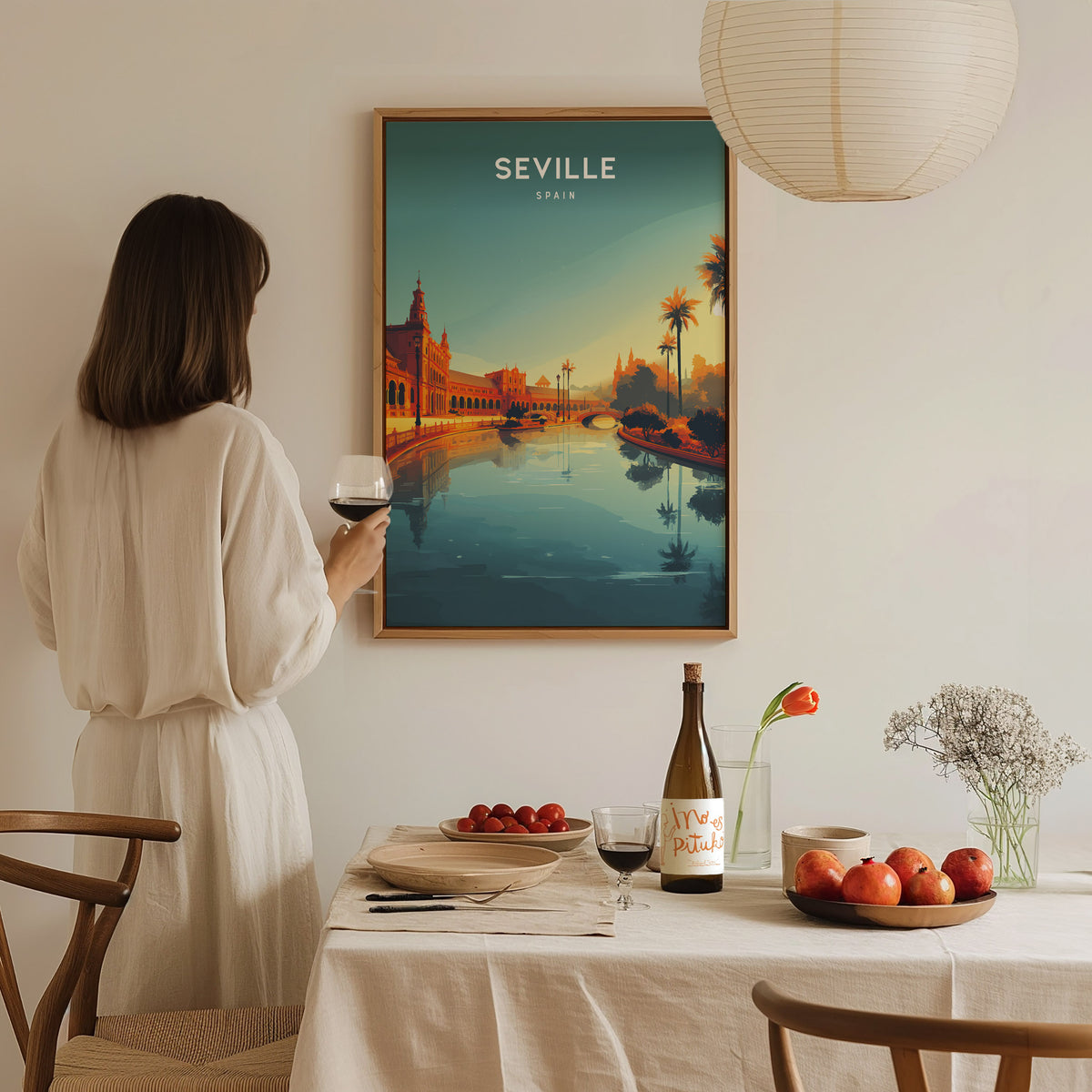 A woman in a light-colored dress holds a glass of wine while standing near a dining table. The table is set with a bottle of wine, a plate of apples, and a vase of flowers.