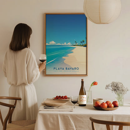A woman in a white outfit stands near a dining table, holding a glass of wine, while admiring a framed poster of Playa Bavaro. The table is set with a bottle of wine, a plate of fruits, and decorative elements in a cozy, well-lit room.