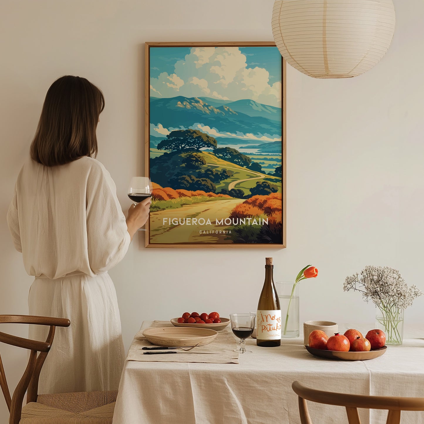 A woman in a light-colored dress stands beside a dining table, gazing at a framed poster of Figueroa Mountain. The table is set with a bottle of wine, a plate of apples, and a vase with flowers, creating a cozy, inviting atmosphere.