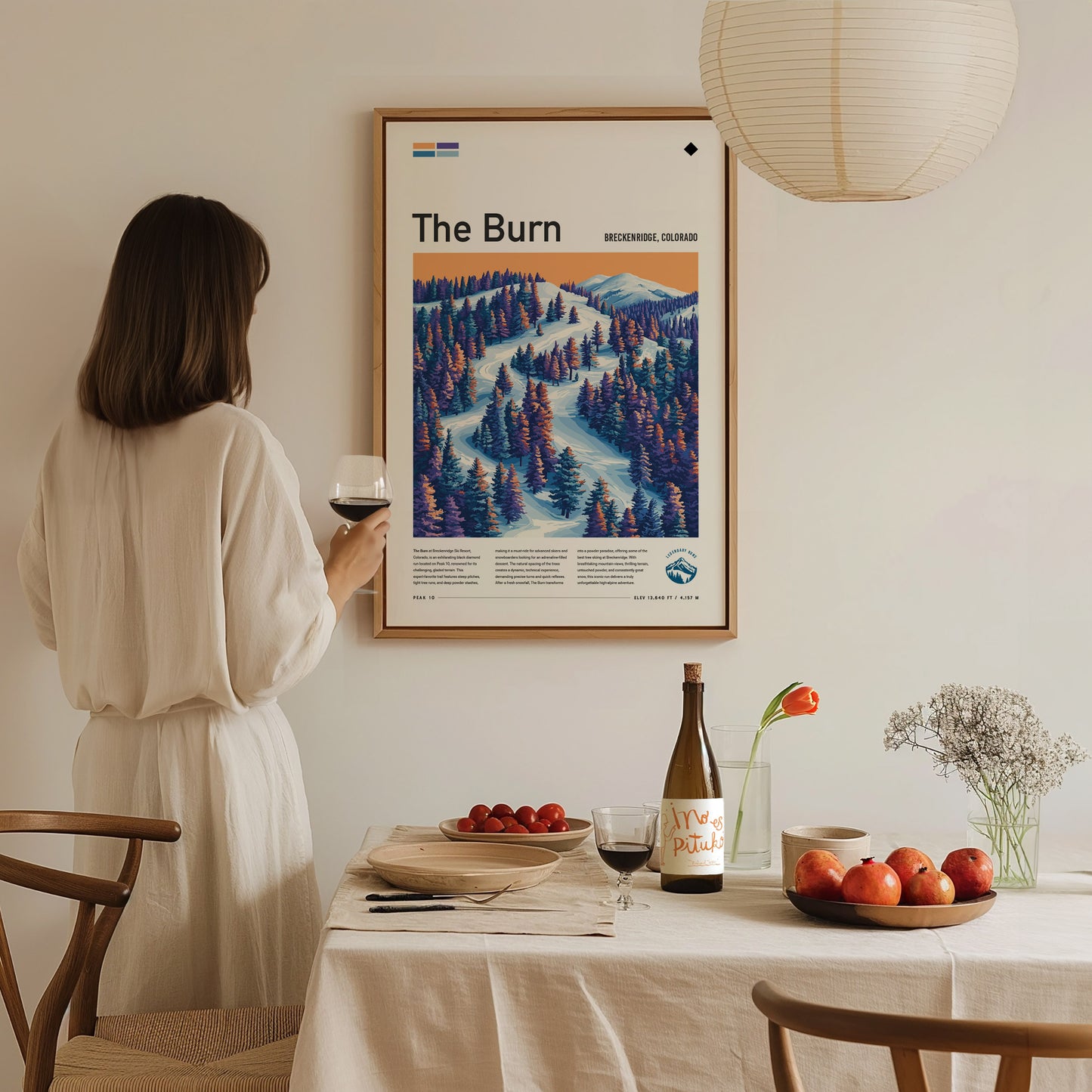 A woman in a white dress holds a glass of wine while admiring a framed poster titled The Burn, depicting a snowy ski run with pine trees.