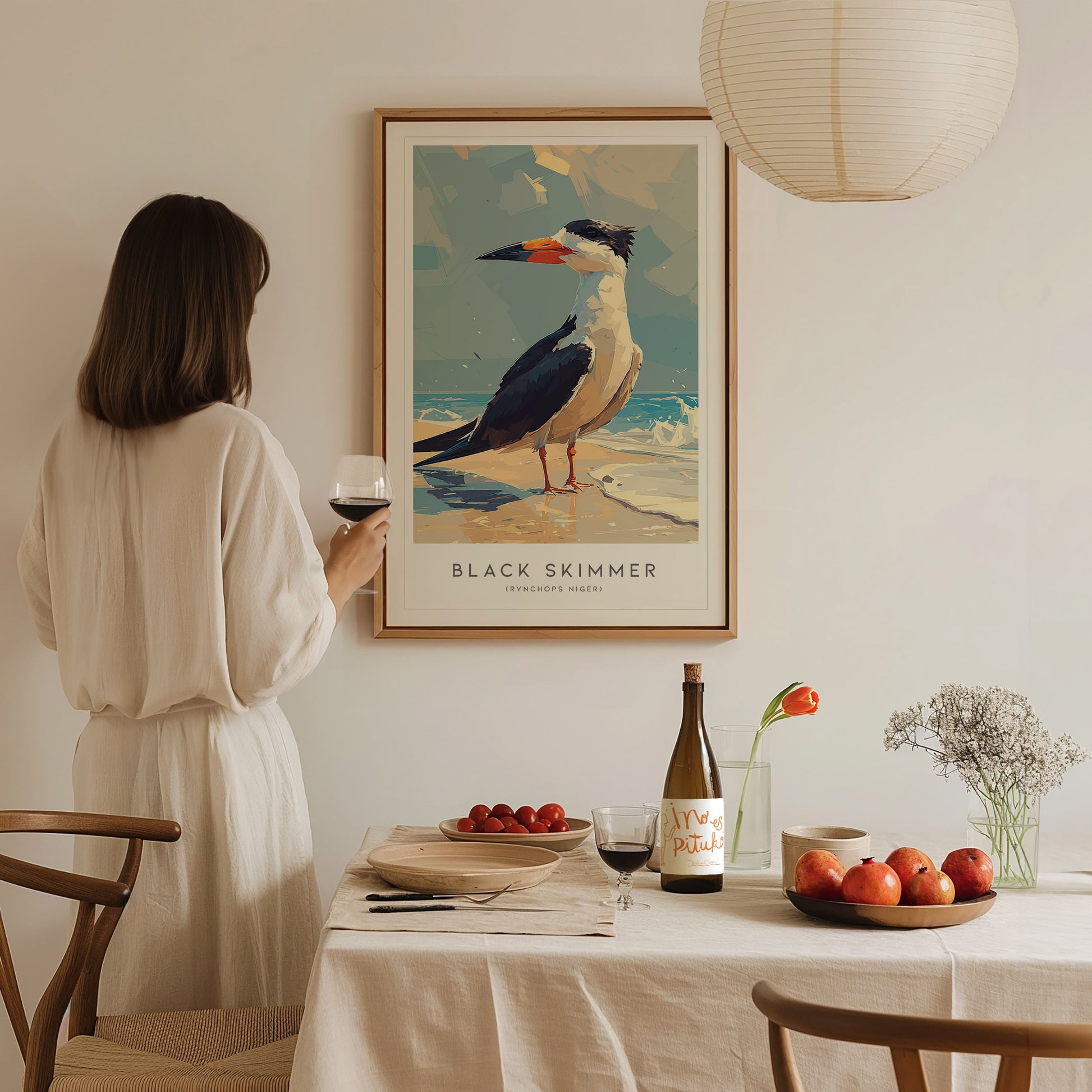 A woman in a white outfit stands beside a dining table, admiring a framed poster of a Black Skimmer on the wall. The table is set with a bottle of wine, a plate of apples, and a vase of flowers, creating a cozy atmosphere.
