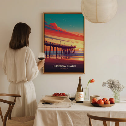 A woman in a white dress holds a wine glass, admiring a Hermosa Beach poster depicting a vibrant sunset pier scene.