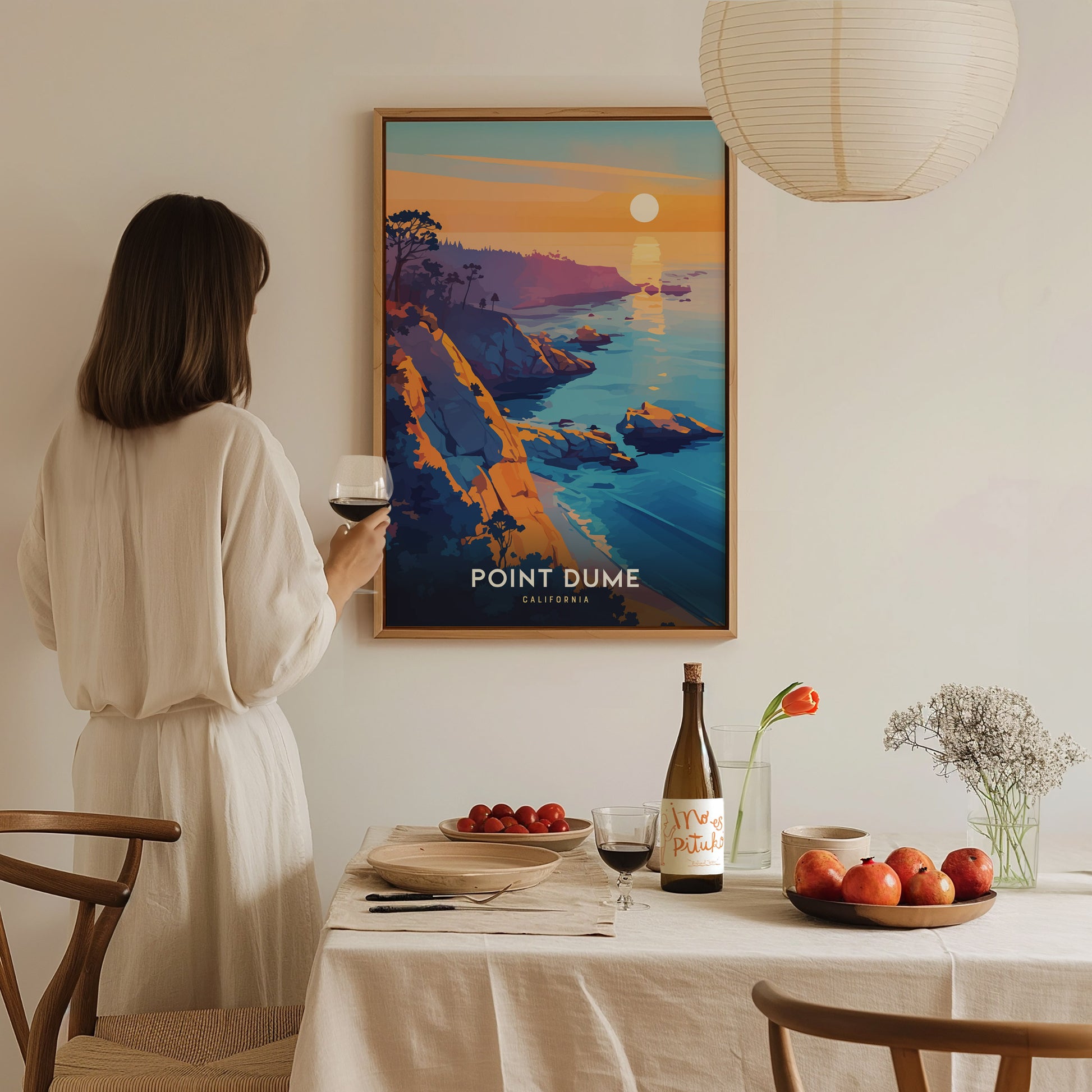 A woman in a white dress holds a glass of wine while admiring a Point Dume poster. The table is set with fruit, a wine bottle, and a vase with.