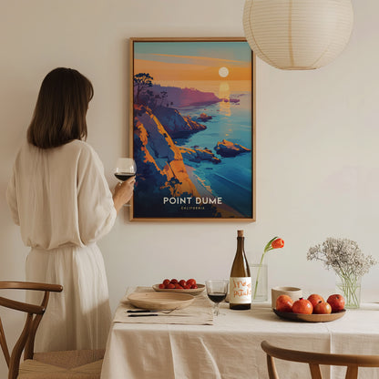 A woman in a white dress holds a glass of wine while admiring a Point Dume poster. The table is set with fruit, a wine bottle, and a vase with.