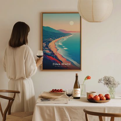 A woman in a white outfit holds a glass of wine while standing next to a framed poster of Zuma Beach, showcasing a colorful coastal landscape at sunset.