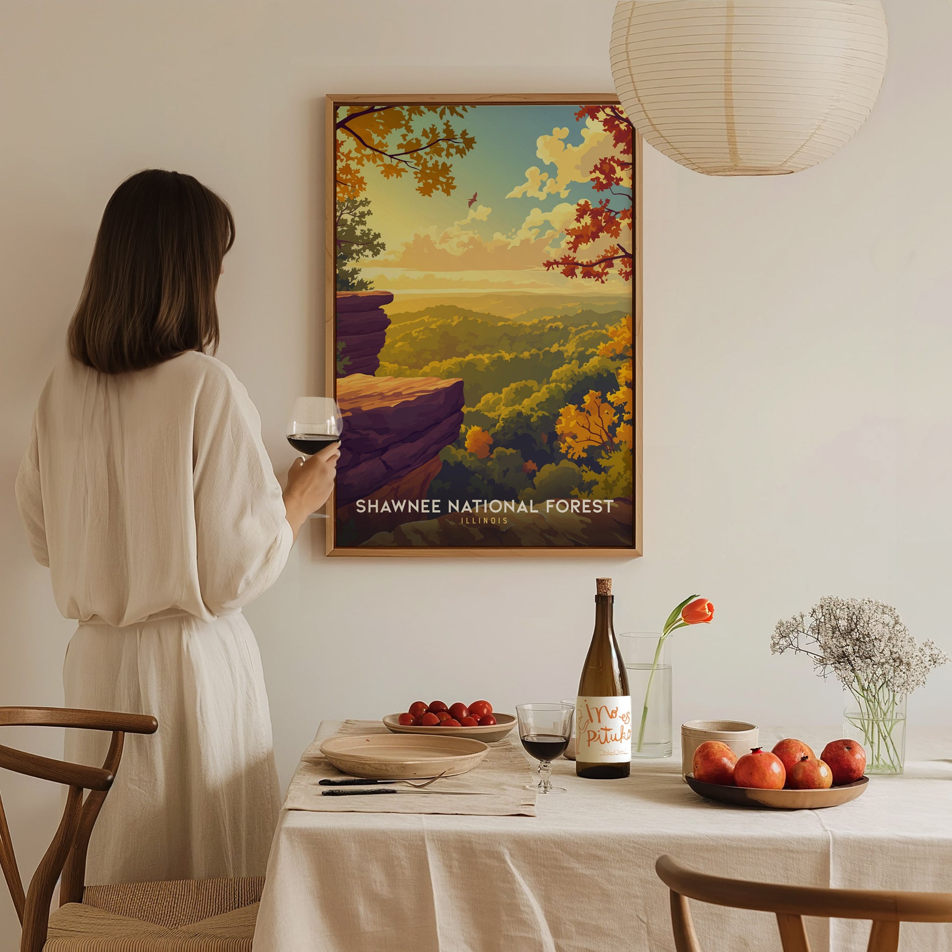 A woman in a white dress stands beside a dining table, holding a glass of wine, while admiring a colorful poster of Shawnee National Forest. The table is set with a bottle of wine, fruit, and a vase of flowers, creating a cozy atmosphere.