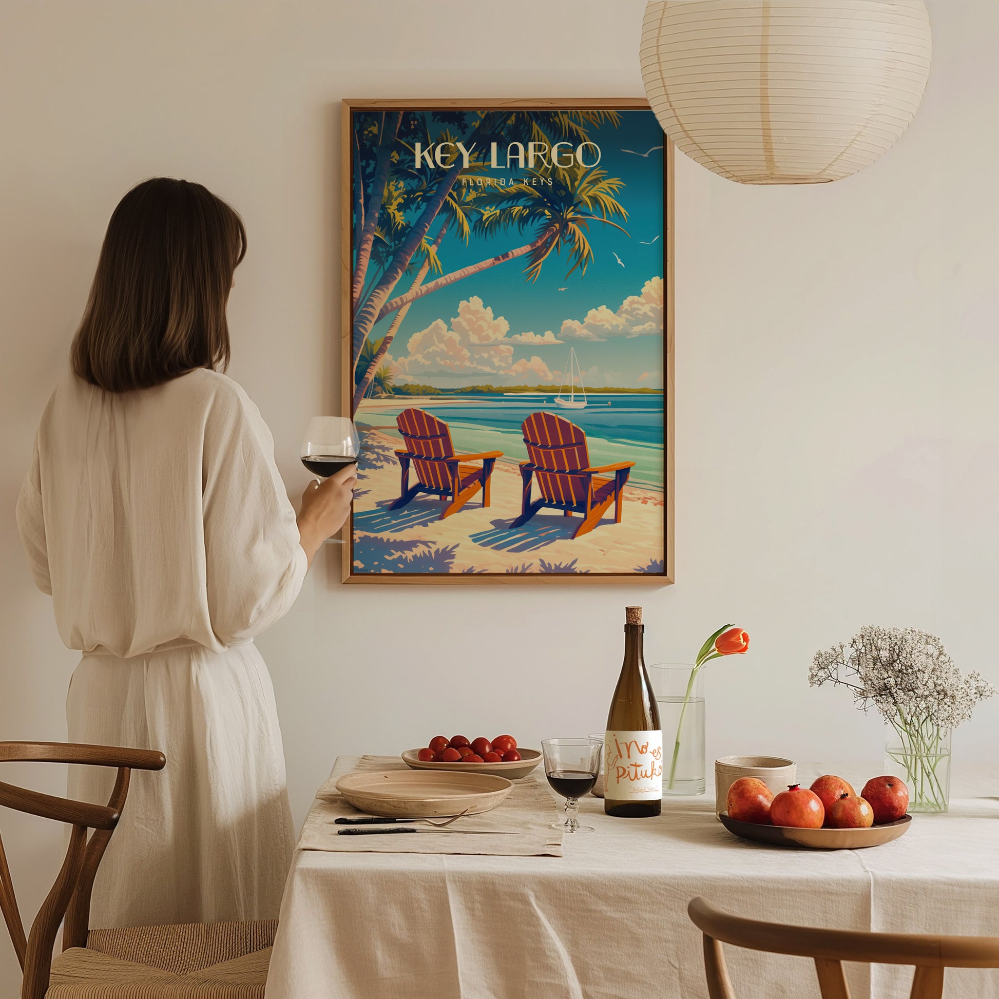 A woman in a white dress holds a glass of wine while admiring a Key Largo poster featuring beach chairs and palm trees.