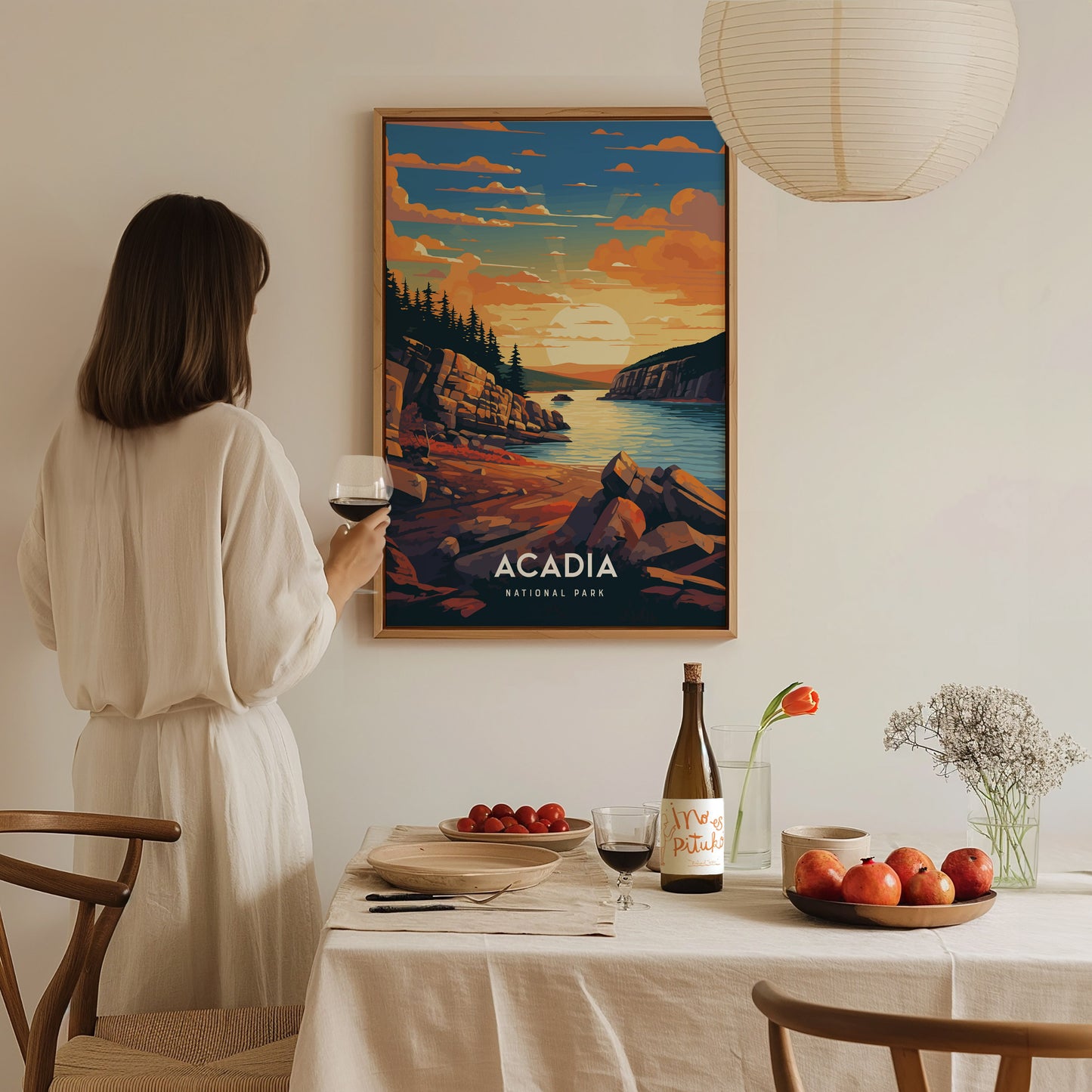A woman in a white dress admires an Acadia National Park poster depicting a coastal sunset. A table with wine, fruit, and flowers is set in the foreground.