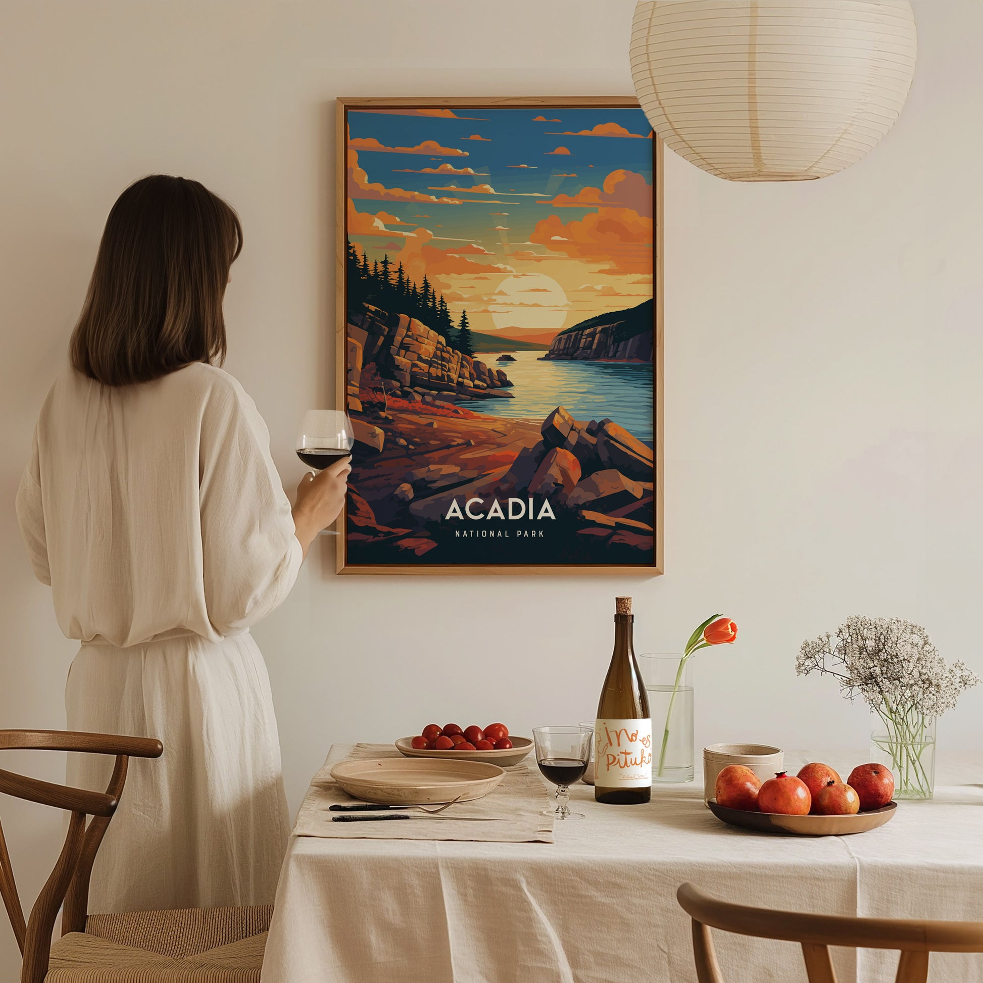 A woman in a white dress admires an Acadia National Park poster depicting a coastal sunset. A table with wine, fruit, and flowers is set in the foreground.