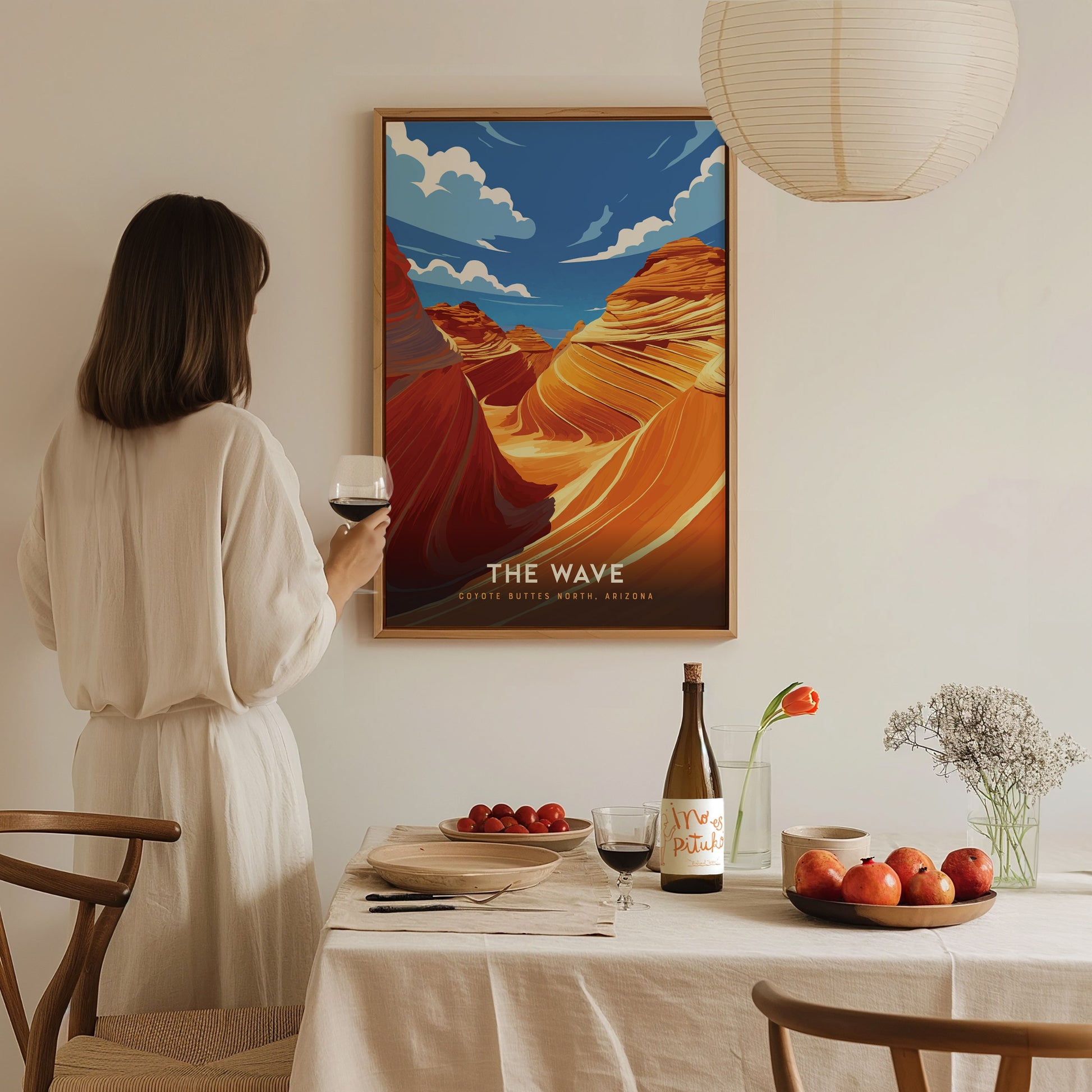 A woman holding a wine glass admires a framed poster of The Wave, featuring vibrant desert formations. The scene includes a dining table with fruits and wine.