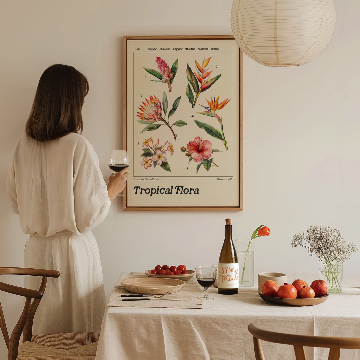 A woman in a white dress admires a framed botanical poster of tropical flowers in a dining room setting with wine, apples, and a tulip on the table.