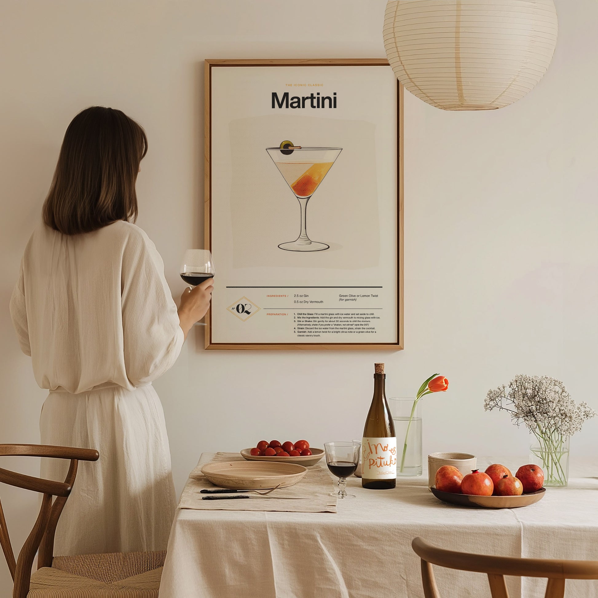 A woman in a white dress stands by a table, holding a wine glass, looking at a framed martini cocktail poster.