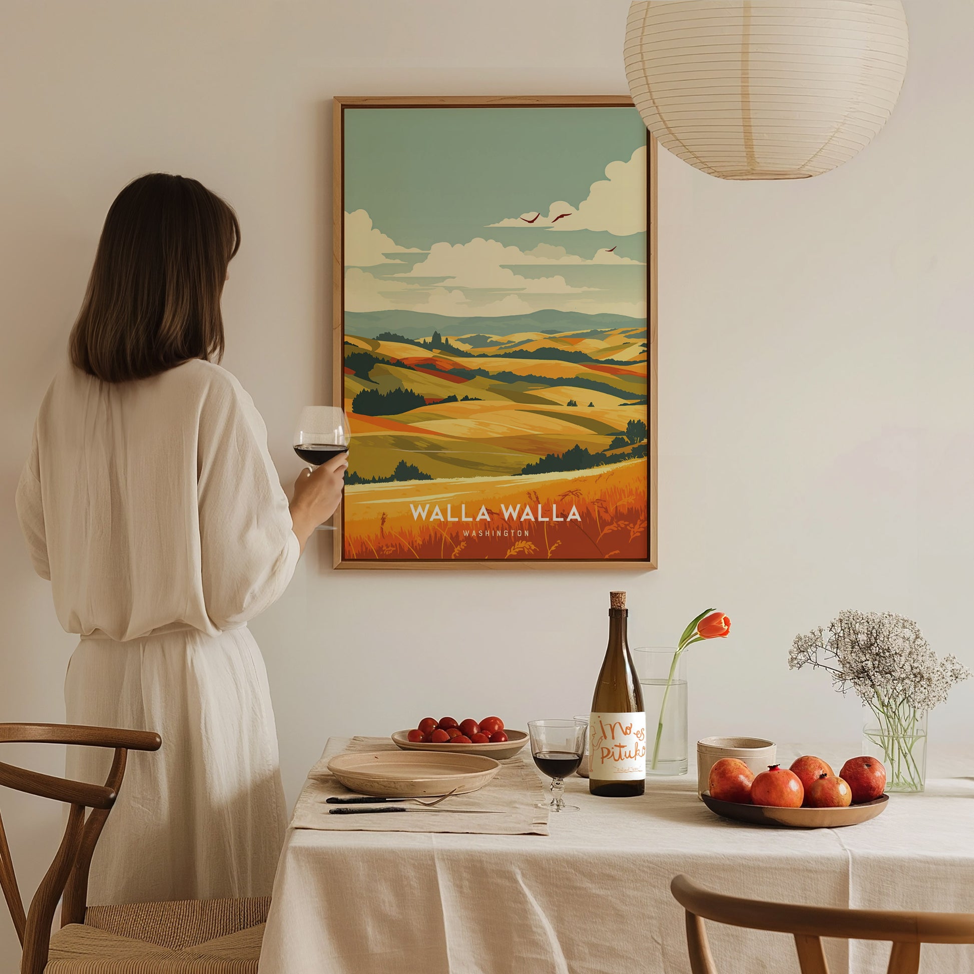 A woman in a white dress holds a glass of red wine, gazing at a colorful Walla Walla landscape poster. A table with fruit and wine is in the foreground.